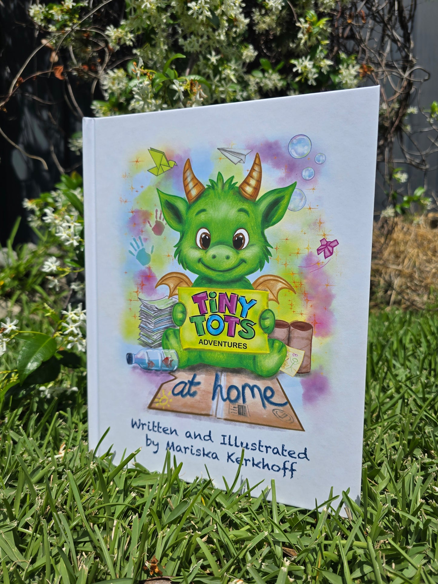 Children's book titled 'Tiny Tots Adventures at Home' with a green dragon illustration on a grassy background.