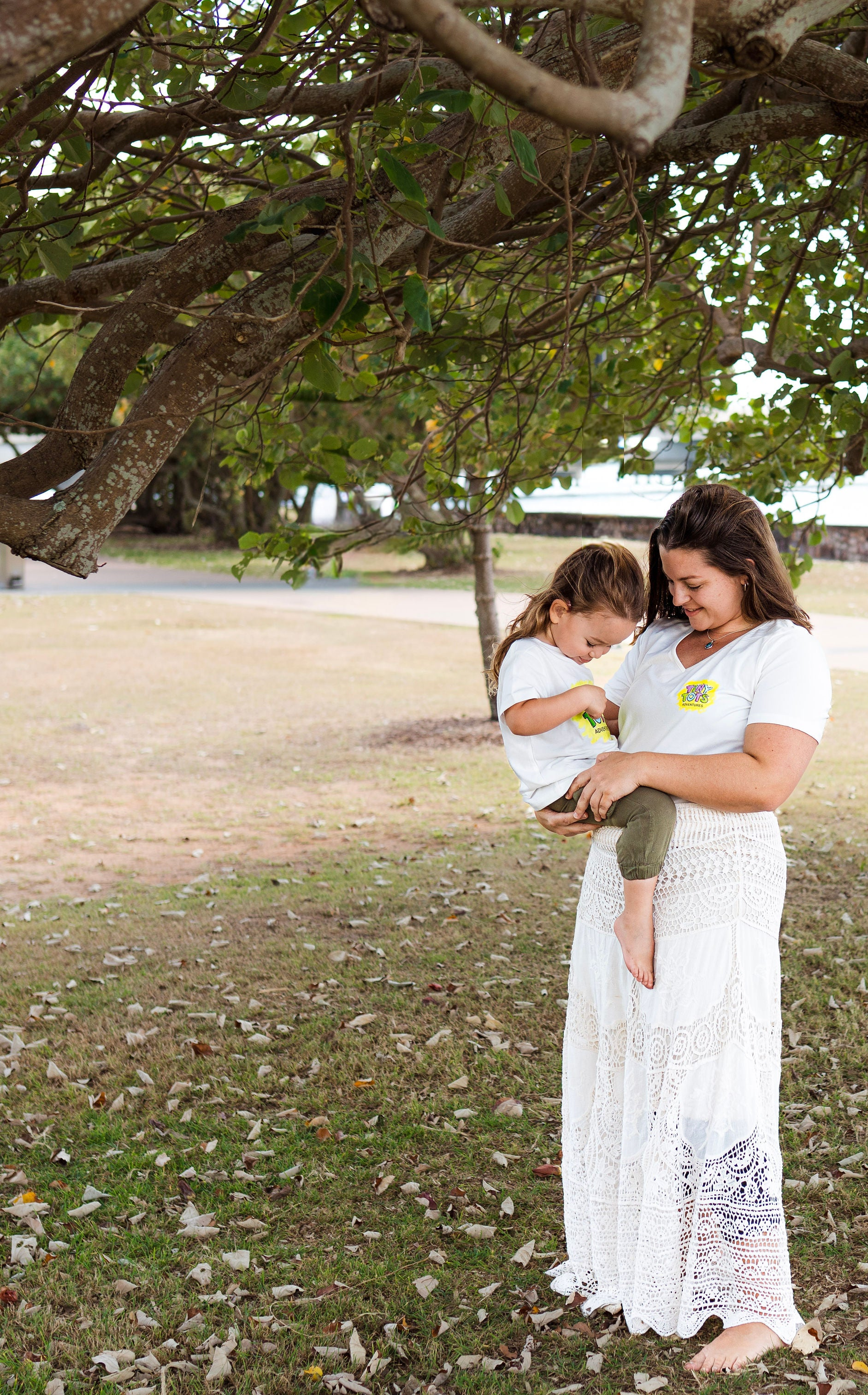 Woman and child in white dresses standing under a tree in a park.