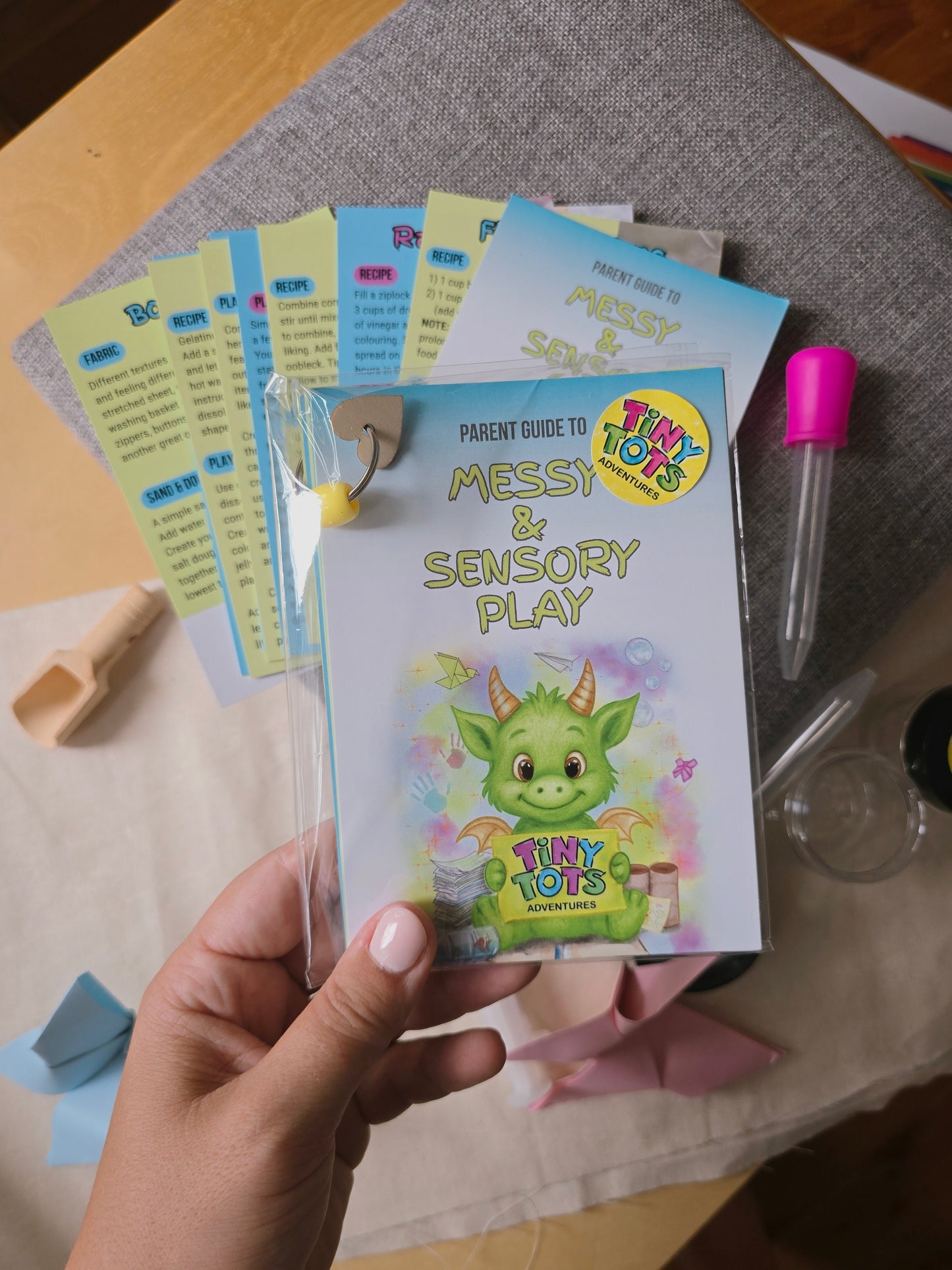 hand holding wrapped pack of parent guide to messy and sensory play on a background of sensory play materials