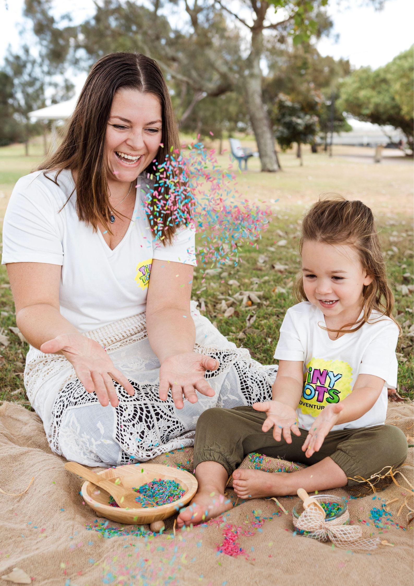 Woman and young girl sitting on a blanket outdoors, engaging in an activity with colorful items.